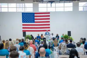 A speaker addresses an audience indoors before a large American flag, as Family Medicine Residents from the New Class sit onstage.