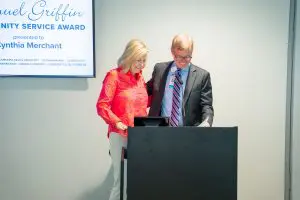 A woman and a man stand together behind a podium, smiling. A screen reads Lemuel Griffin Community Service Award presented to Cynthia Merchant.