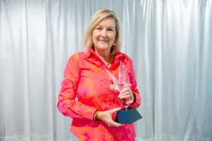 A smiling woman in a bright pink and orange blouse proudly holds the Lemuel Griffin Community Service Award before a pale curtain.