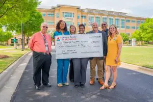 Seven people stand outside a large building, smiling and holding a $50,000 check to Phoebe Hospice, with trees in the background.