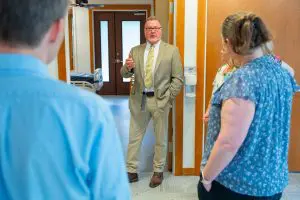 A man in a tan suit speaks to three people near a hospital bed at Phoebe Hospice. The others listen attentively, backs to camera.