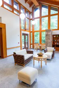 A modern lounge at Phoebe Hospice with armchairs, sofa, wooden tables, stone pillar, high ceilings, windows, and garden views.