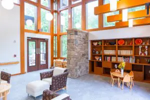 A bright, modern lounge at Phoebe Hospice with large windows, wood accents, bookshelves, and cozy seating filled with natural light.