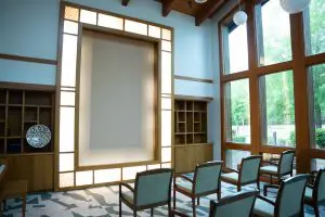 A small, modern chapel at the hospice features wooden chairs, illuminated panel, wood accents, shelves, and large sunlit windows.