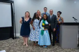 Six adults and a young woman in graduation attire stand smiling indoors; the graduate holds a diploma. A podium, screen behind them.