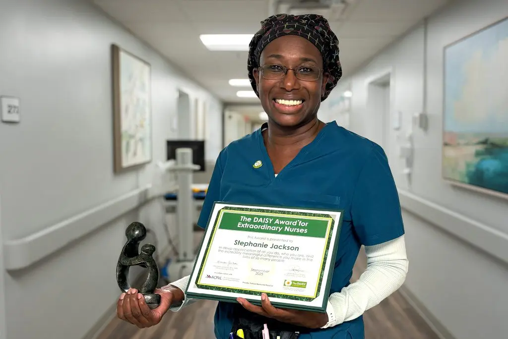 A smiling PPMH nurse stands in a hospital hallway holding a DAISY Award certificate and statue, celebrating her achievement.