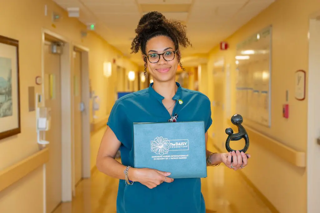 A nurse in glasses and scrubs stands in a hallway holding a DAISY Award certificate folder and black trophy, smiling.