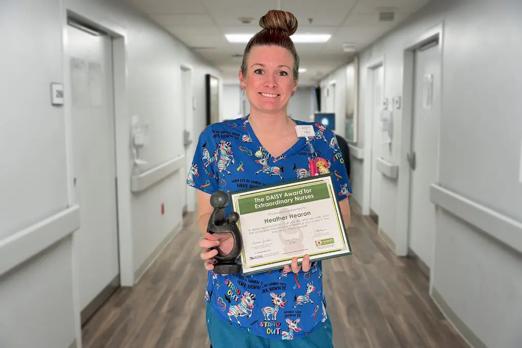 A nurse in blue, cartoon-themed scrubs stands in a hospital hallway, smiling and holding a certificate and sculpture after receiving the DAISY Award.