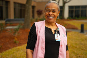 Barbara Banks, smiling in a pink vest and name badge stands outside near a building and lawn, celebrating Volunteer Appreciation Week.