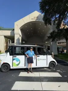 Charles Proctor in a blue shirt stands by a shuttle van labeled "Phoebe," celebrating volunteers outside Main Entrance on a sunny day.