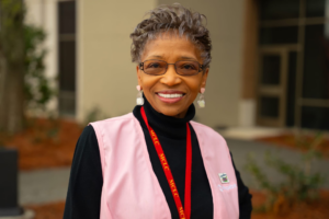 Jonelle Cooks, smiling in glasses and a pink vest stands outdoors, wearing a name badge and red lanyard for Phoebe’s Mission volunteers.