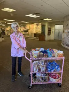 Millie Proctor in a pink jacket stands by a snack cart during a Phoebe’s Mission volunteer appreciation event in the lobby area.