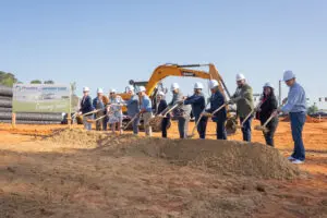 A group in hard hats and business attire hold shovels at a Lee County groundbreaking; an excavator and Phoebe Urgent Care sign behind.