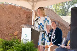 A woman in a white dress with blue flowers speaks at a Phoebe Urgent Care podium, with two men seated and dirt behind her.
