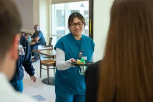 A woman in teal scrubs hands a dessert plate to a person celebrating a bariatric surgery milestone in a bright cafeteria.