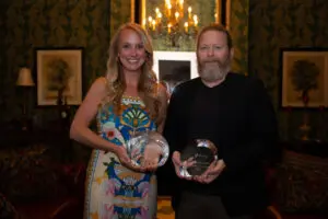 A woman in a colorful dress and a man in a dark jacket, honored as Outstanding Physician, hold glass awards and smile while standing in a warmly lit room with patterned wallpaper, framed art, and a chandelier in the background.