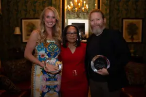 Three people pose indoors at an event. The woman on the left wears a colorful dress and holds the APP of the Year award. The woman in the center wears a red dress. The bearded man on the right holds his Outstanding Physician award amid elegant décor.