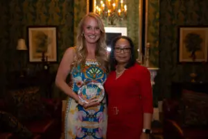 Two women stand side by side in a warmly lit, elegant room. The woman on the left holds an APP of the Year glass award and wears a colorful dress, while the woman on the right wears a red outfit beneath a chandelier.