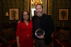 A woman in a red dress, stands next to a man in a black jacket holding the APP of the Year glass award, both smiling in an elegant, warmly lit room with patterned wallpaper and framed art.