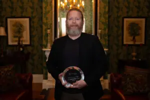 A man with a beard, wearing a black blazer, stands indoors holding an Outstanding Physician award. Behind him are ornate wallpaper, framed art, a fireplace, and a lit chandelier in Phoebe’s elegant and formal setting.