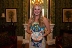 A smiling woman with long blonde hair holds a clear APP of the Year award indoors, with chandelier, white mantle, and green wallpaper.