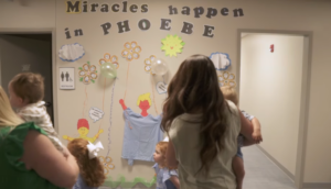 Two women and children read a sign that says “Miracles happen in PHOEBE” at a Children’s Miracle Network celebration.