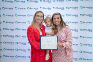 Two women holding a certificate and a child smile before a Phoebe Sumter step-and-repeat backdrop.