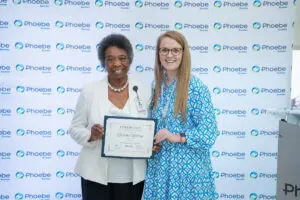 Two women smiling, one nurse holding a DAISY Award certificate, in front of a Phoebe Sumter logo backdrop.