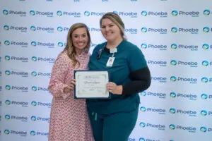 Two women smiling, one nurse in scrubs holding a certificate, in front of the Phoebe Sumter logo backdrop.