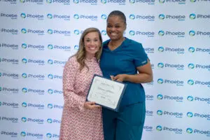 Two women smile as one, a nurse from Phoebe, holds a certificate before a Phoebe Sumter backdrop.