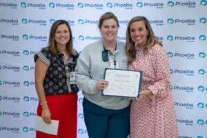 Three women stand together smiling; a nurse holds a certificate in front of a Phoebe Sumter branded backdrop.