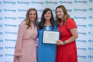 Three women smiling; a nurse in blue holds a certificate in front of a Phoebe Sumter logo step-and-repeat backdrop.