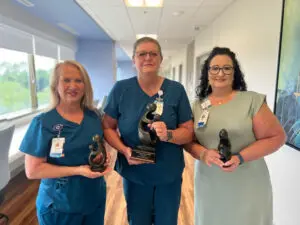 Three women indoors, two in blue scrubs, smiling and holding DAISY Award trophies for compassionate care.