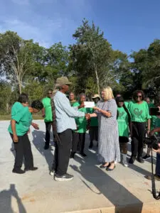 A man and woman shake hands outdoors as people in green shirts smile around them on a sunny day.