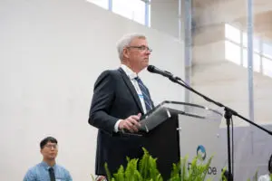 An older man in a suit speaks at a podium for Dream Community Kitchen, with another man seated in the background.