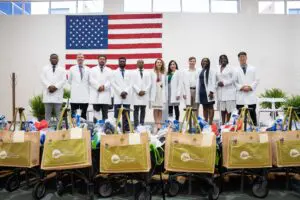 A group in white coats from Phoebe Family Residency stands before the American flag and carts filled with supplies.
