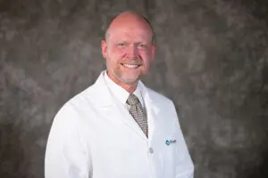 Smiling electrophysiologist, Dr. James Coman, in a white lab coat and patterned tie, standing against a gray mottled background.