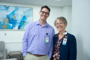 Two smiling people wearing PPMH name badges stand together in an office with abstract blue art displayed on the wall behind them.