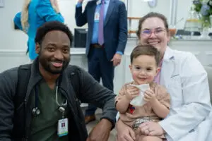A smiling doctor, nurse, and young child pose together in a cheerful Family Medicine Residency Clinic setting.