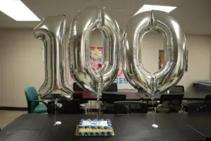 Large silver "100" balloons and a cake suggest a centennial celebration, perhaps linked to an electrophysiologist or pacemaker.