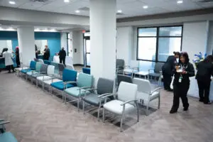 A modern waiting room at Phoebe Primary Care, featuring empty chairs and groups of people talking near windows and walls.