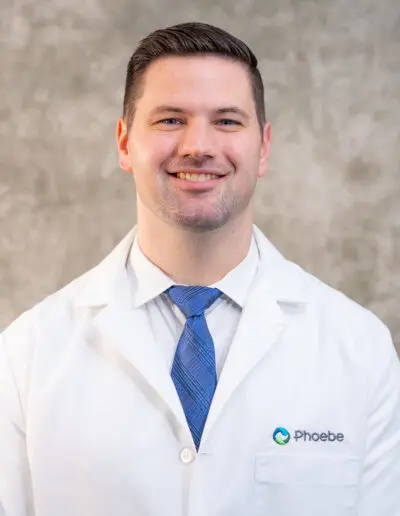 A man in a white lab coat with a Phoebe logo smiles, representing the Family Medicine Residency.