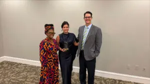 Three people stand together smiling. The woman in the center holds an award at Family Medicine Residency Graduation Ceremony.