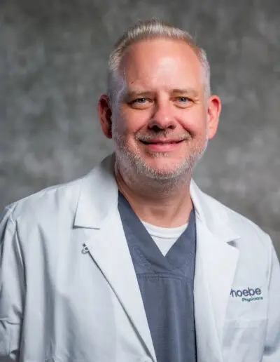 A smiling man in a white lab coat and gray scrubs stands before a gray backdrop, representing Family Medicine Residency.
