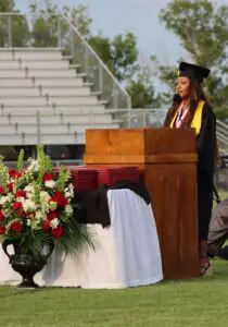 A graduate in cap and gown, who once credited the NICU with saving her life, speaks at a podium on a field.