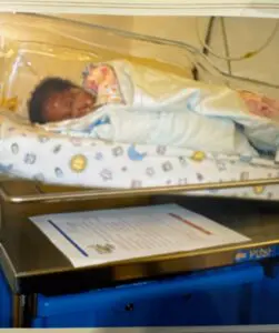 Newborn baby wrapped in blankets, resting in a hospital bassinet under a clear cover with neonatal care paperwork underneath.