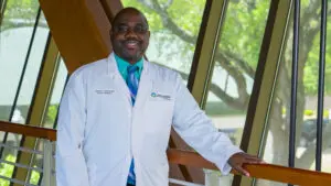 Despite Confusion over Vaccine Guidance Phoebe Says the Medical Recommendation Hasn’t Changed 3 Smiling doctor in a white coat stands by a railing indoors, ready to offer expert vaccine guidance.