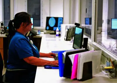 A nurse in blue scrubs types at a computer in a Phoebe organization medical office with paperwork and monitors nearby.