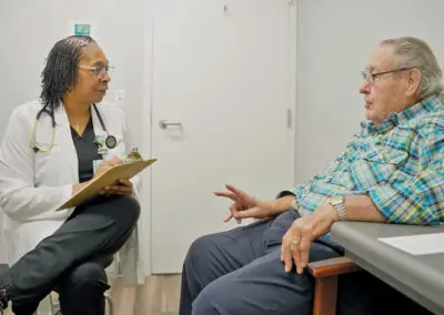 A doctor with a clipboard speaks with an older man in a rural clinic exam room. Both sit and converse attentively.