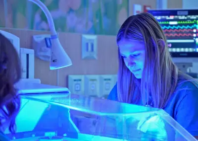 Two healthcare workers from Phoebe care for an infant in an incubator under blue light, monitors glowing in the background.